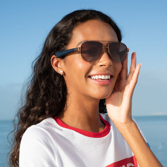 A woman with long curly hair smiles while wearing Nautica Admiral polarized aviator glasses and a white T-shirt with red trim, standing outdoors near the ocean on a sunny day.