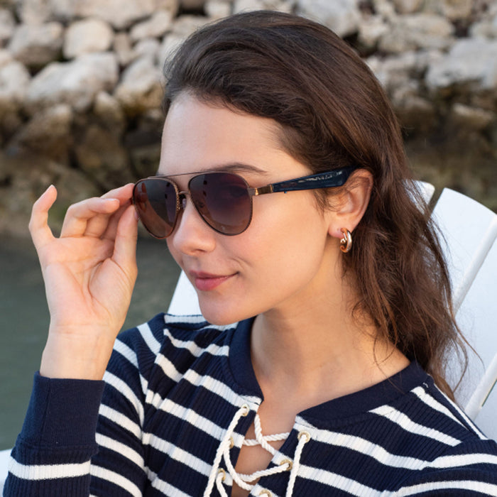 A woman in Nautica Admiral polarized aviator sunglasses and a navy and white striped shirt sits on a white chair outdoors by water and rocks, smiling slightly as she touches her Nautica sunglasses.