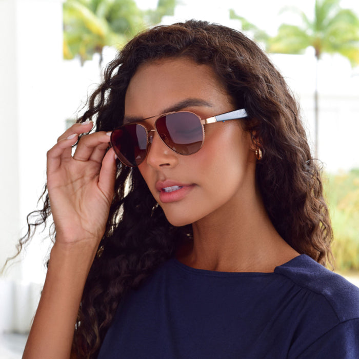 A woman with long curly hair wears Nautica Admiral smart audio sunglasses and a navy blue top, looking to the side and adjusting her glasses. Palm trees and blurred outdoor scenery are in the background.