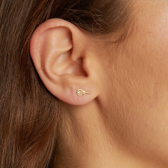 Model. Close-up of an ear wearing a gold earring with a tennis racket design.