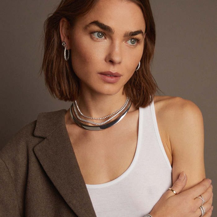 Model. Woman wearing a brown blazer and white top with layered necklaces against a neutral background
