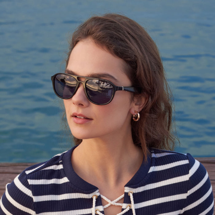 A woman with wavy brown hair wearing Nautica Commander polarized aviator sunglasses and a navy-and-white striped top sits outdoors by the water, gazing slightly to the side.