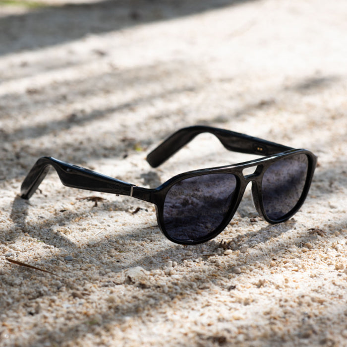 The Nautica Commander polarized aviator sunglasses with dark lenses rest on light-colored sand, with tree shadows cast across the ground.
