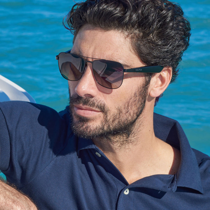 A man with dark curly hair and a beard wears Nautica Corsair polarized aviator sunglasses and a navy blue polo shirt, sitting outdoors near bright blue water on a sunny day.