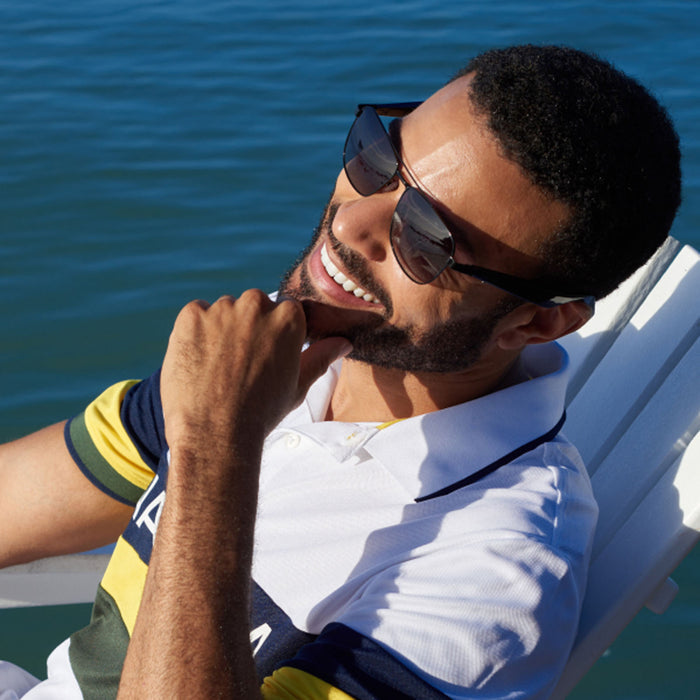 A man in Nautica's Corsair smart sunglasses and a striped polo shirt smiles as he relaxes on a white chair by the water in the bright sun.