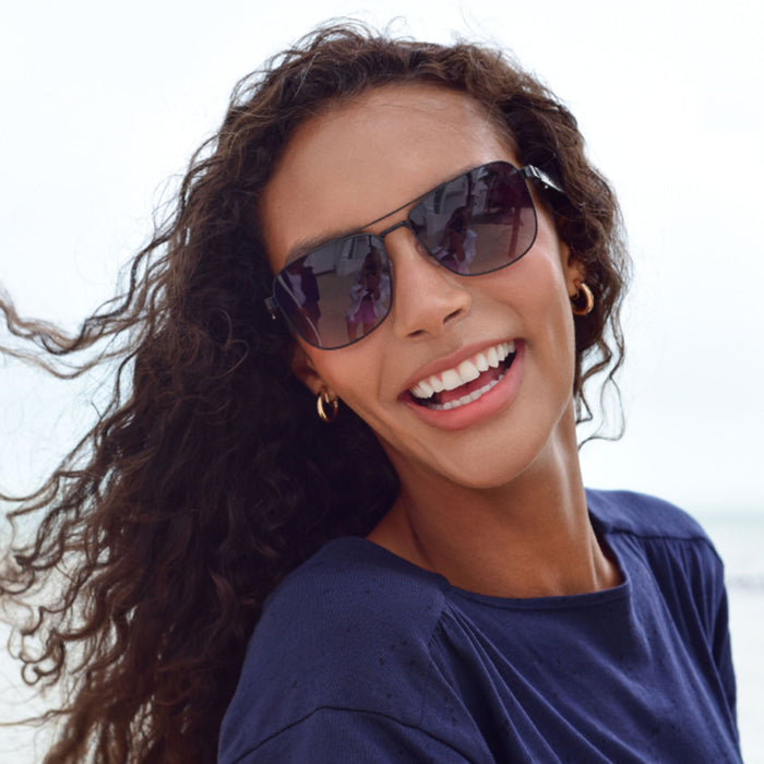 A woman with curly hair smiles outdoors on a beach, wearing Nautica Corsair polarized aviator sunglasses and a navy blue top.