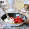 Coupelle en porcelaine blanche, intérieur noir et bordure or, contenant du fromage blanc et des groseilles.