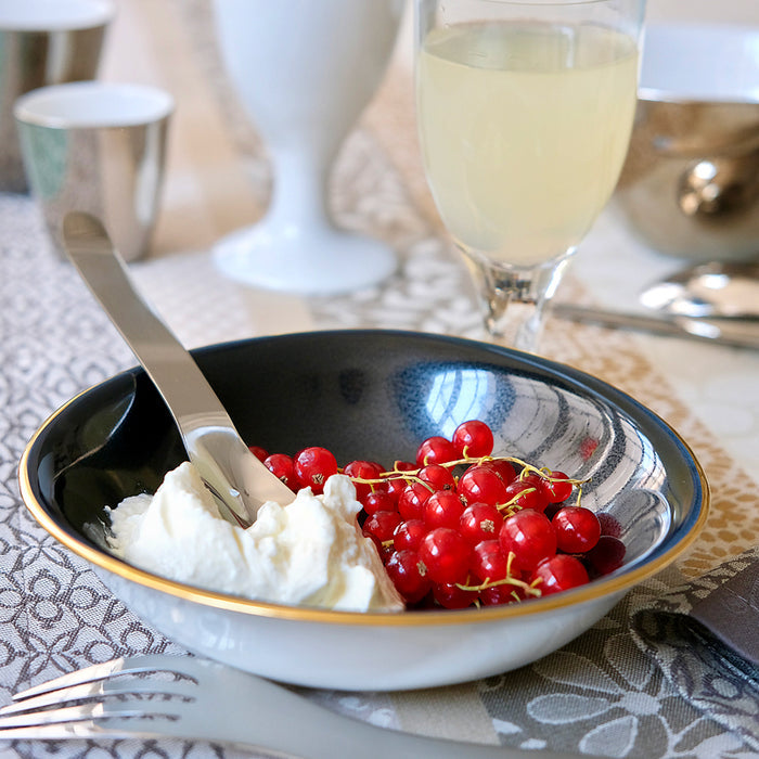 Coupelle en porcelaine blanche, intérieur noir et bordure or, contenant du fromage blanc et des groseilles.