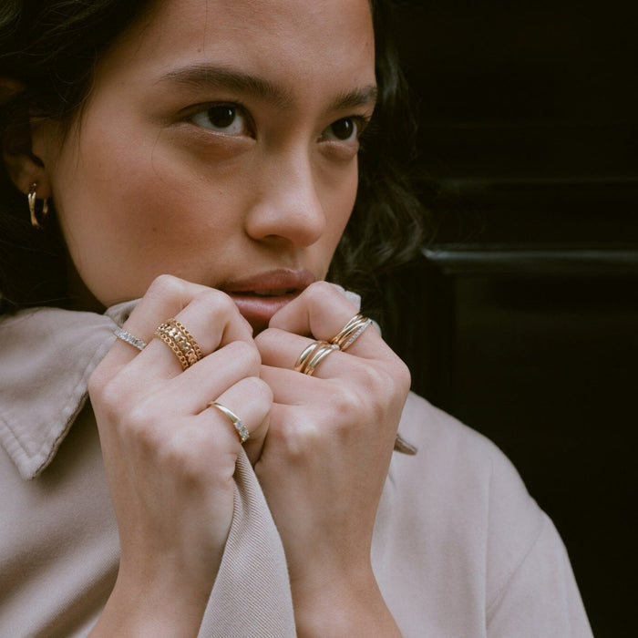 Lifestyle Model Close-up of a woman resting her hands near her lips, wearing a mix of gold rings—including a thick chain-link style and a classic gold and diamond crossover ring—paired with a small gold hoop earring
