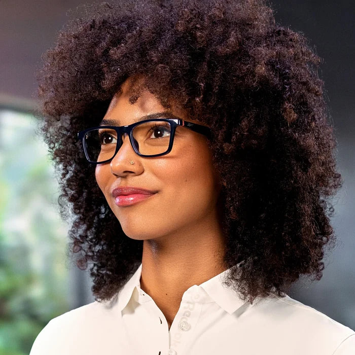 A person with curly hair wears Reebok Dash dark-rimmed smart glasses and a white collared shirt, smiling softly while glancing to the side against a blurred green background.