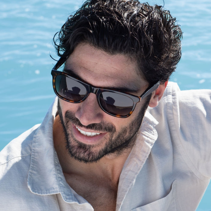 A dark-haired, bearded man in a light shirt smiles by the water on a sunny day, enjoying his Nautica Dockside Bluetooth audio glasses.