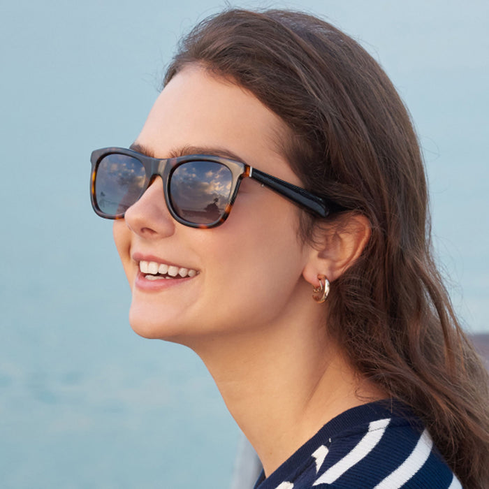 A woman with long brown hair smiles outdoors near the water, wearing Nautica's Dockside smart sunglasses, gold hoop earrings, and a navy striped top, their lenses reflecting a sky with clouds.