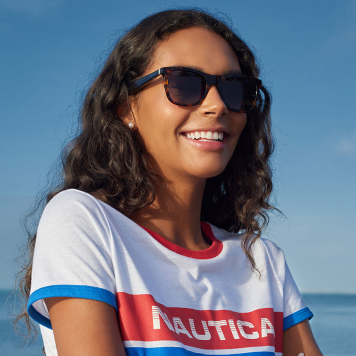 A woman with long curly dark hair smiles outdoors in Nautica Dockside polarized UV400 smart sunglasses and a white T-shirt with red and blue trim, enjoying the sunshine with blue sky and ocean in the background.