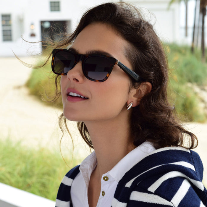 A woman with wavy brown hair wears Nautica Dockside large polarized UV400 sunglasses, silver hoop earrings, and a white collared shirt under a navy and white striped sweater. She stands outdoors near greenery and a white building.