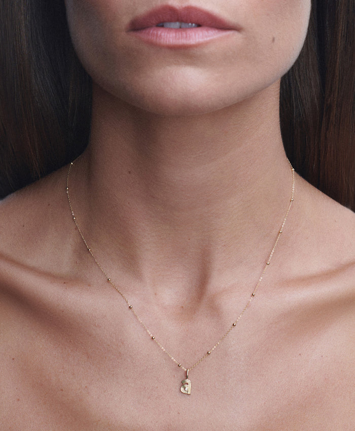 A close-up of a model's neck and upper chest wearing a delicate gold chain necklace with a small gold letter 'a' pendant.