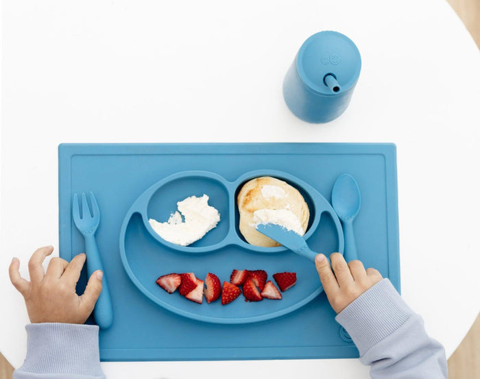 Child using ezpz Happy Utensils Set of 3 in blue together with ezpz mat and cup. Available from tenlittle.com