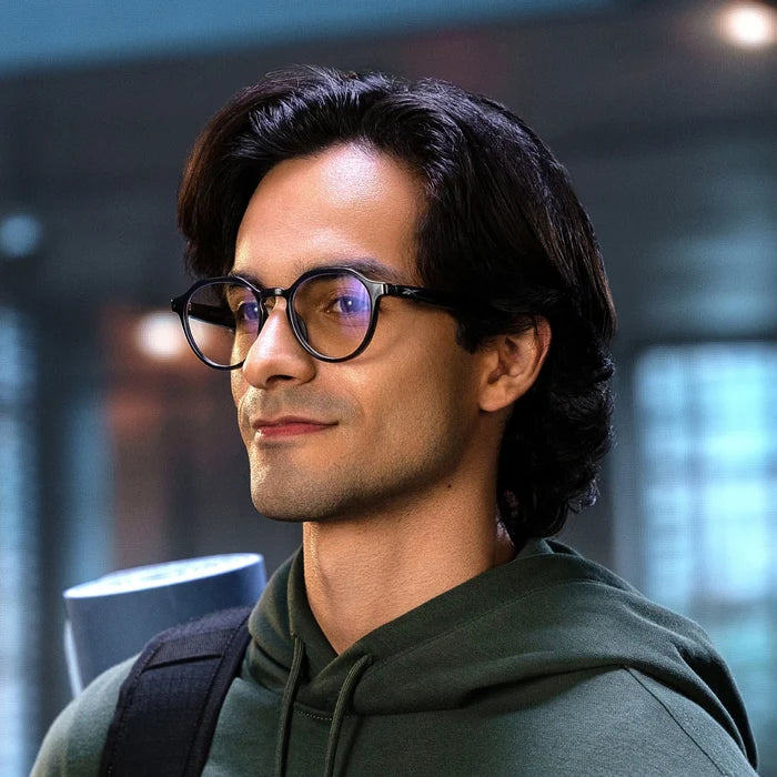 A young man with wavy dark hair and Reebok Jet smart glasses, dressed in a green hoodie, smiles softly while carrying a rolled-up yoga mat over his shoulder in a modern indoor setting.