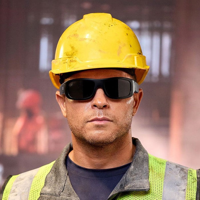 A construction worker in a yellow hard hat, Lucyd Armor | Black Polarized safety glasses (ANSI Z87.1-certified) by Lucyd, and a green reflective vest stands before a blurred industrial background.