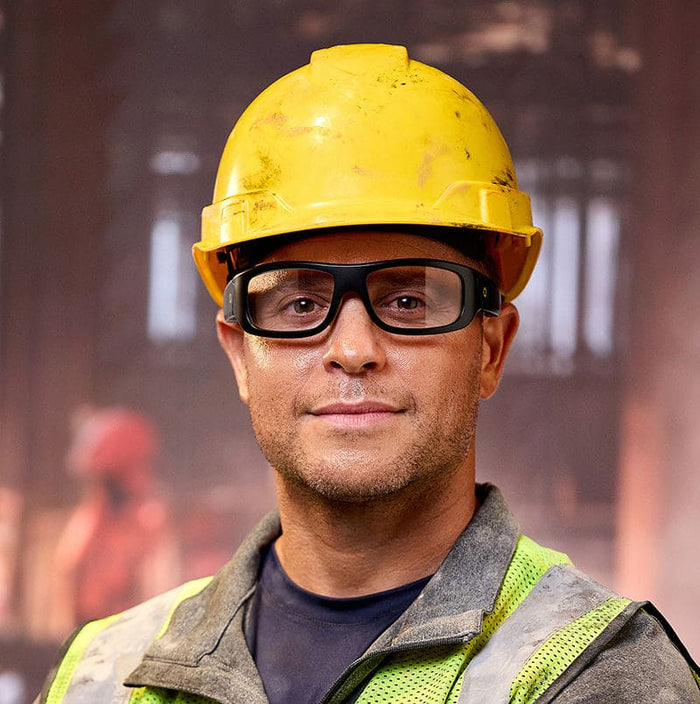 A construction worker in a yellow hard hat, Lucyd Armor | Photochromic Z87.1 safety glasses by Lucyd, and a reflective vest stands indoors with a neutral expression; blurred industrial equipment is visible in the background.