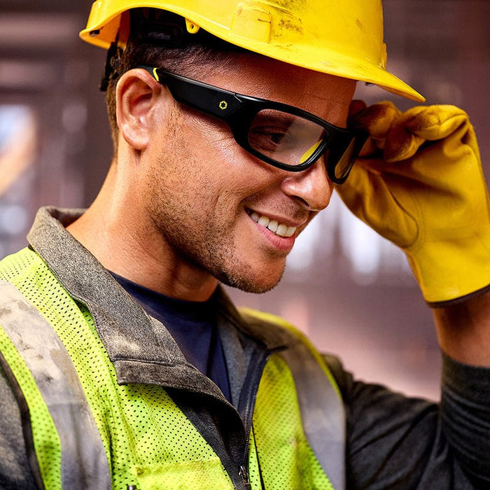 A smiling construction worker in a yellow hard hat, Lucyd Armor | Photochromic safety glasses by Lucyd, yellow gloves, and a reflective vest stands in an indoor work environment.