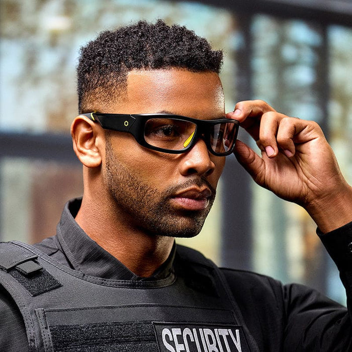 A man in a black security uniform stands indoors, adjusting his Lucyd Armor | Photochromic prescription smart glasses by Lucyd, looking ahead with a focused expression.