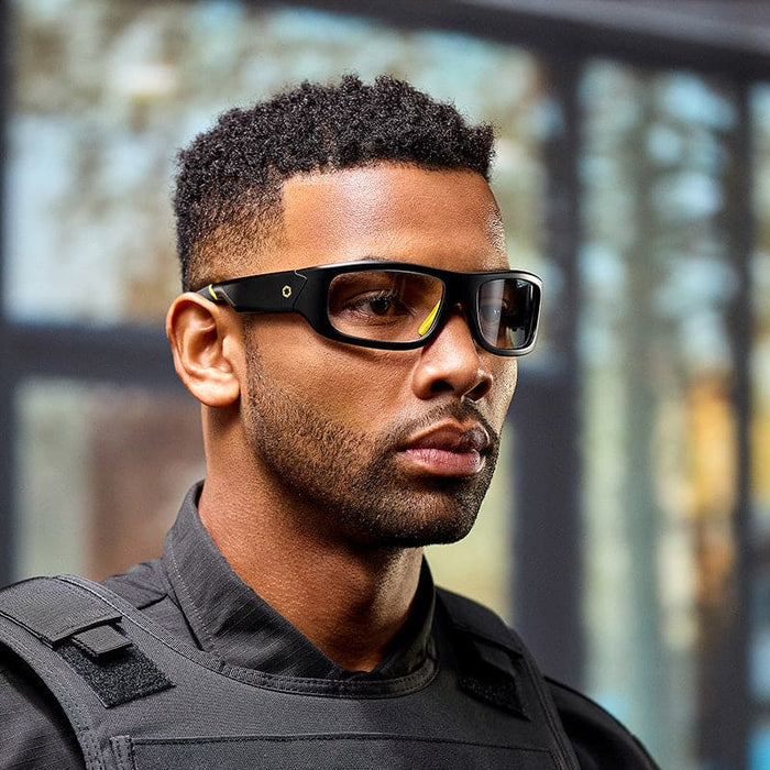 A man indoors wears Lucyd Armor | Photochromic black prescription smart glasses by Lucyd and a tactical vest, looking focused; blurred glass windows and trees are in the background.