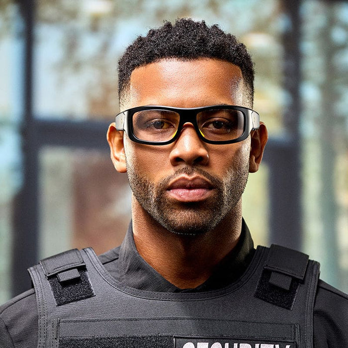 A man wearing Lucyd Armor | Photochromic safety glasses by Lucyd and a security vest stands indoors, looking at the camera with a serious expression. The background is blurred with natural light coming through windows.