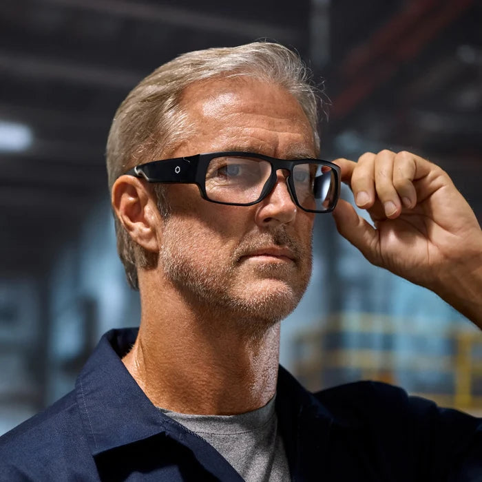 A middle-aged man with short gray hair and a beard wears Lucyd Armor | Vantage Photochromic smart safety glasses by Lucyd and a navy blue shirt, standing indoors in an industrial or warehouse setting while adjusting his glasses.