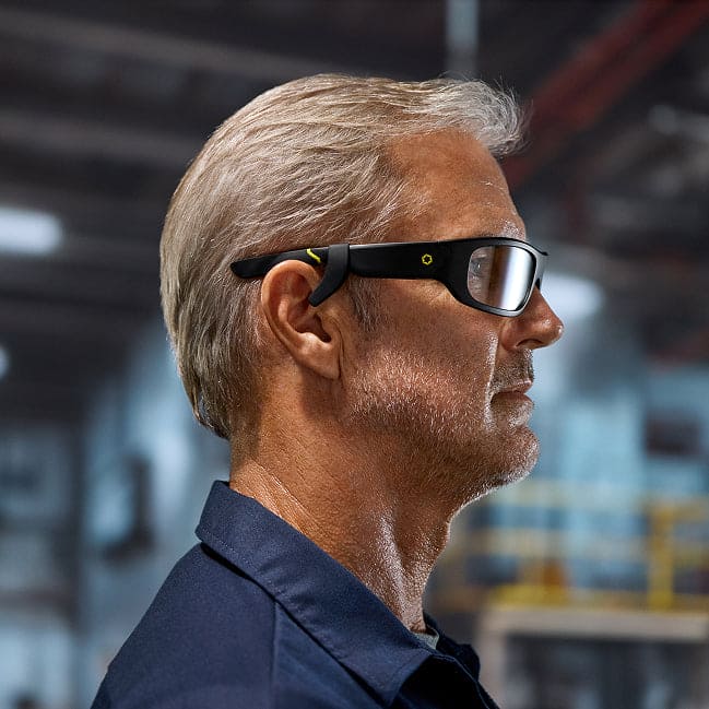 A man with short gray hair and a beard wears Lucyd Boosters for Lucyd Armor & Reebok Sport smart glasses by Lucyd, standing in an industrial or warehouse setting with blurred machinery in the background.