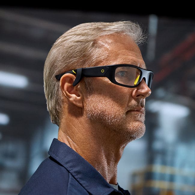 A man with gray hair and a beard wears Lucyd Boosters for Lucyd Armor & Reebok Sport smart glasses by Lucyd and a navy shirt, standing in an industrial setting with blurred machinery in the background.