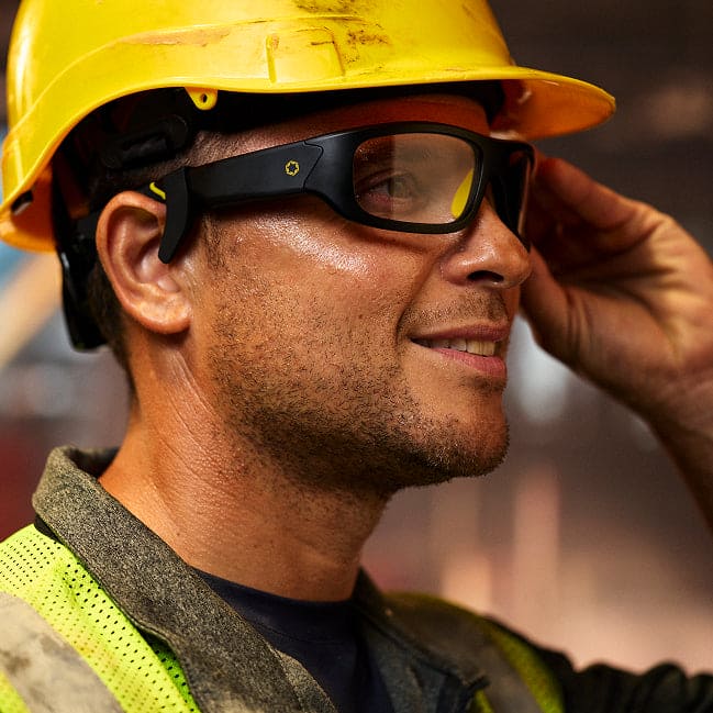 A person in a yellow hard hat, safety glasses, and a reflective vest smiles, touching the side of their Lucyd Boosters for Lucyd Armor & Reebok Sport smart glasses by Lucyd in an industrial setting.