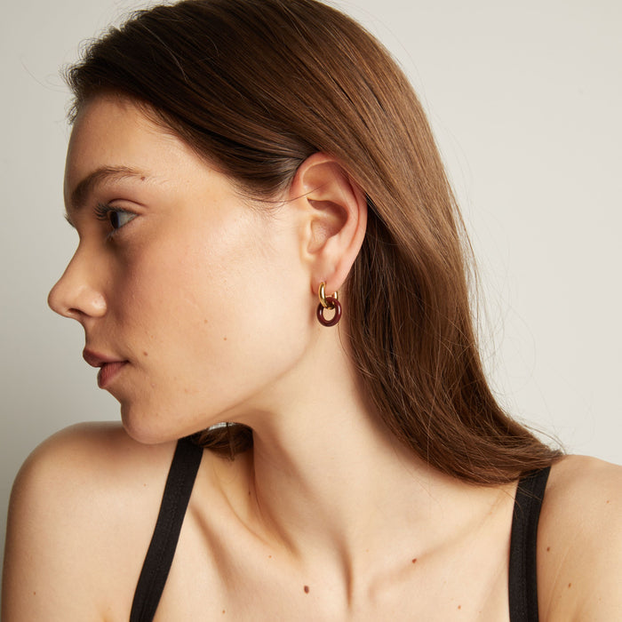 Model. Close up of a person wearing a gold and maroon hoop earring on a neutral background.