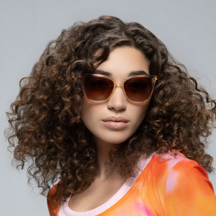 A woman with voluminous curly brown hair wears Lucyd’s Millennia women’s eyewear with brown gradient lenses and a vibrant orange and pink top, posing against a plain light gray background.