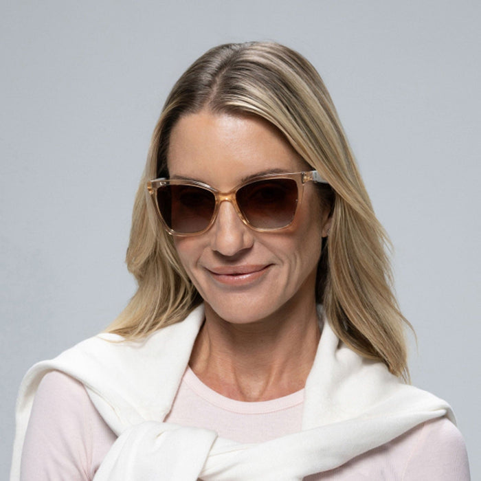 A woman with blonde hair smiles against a plain gray background, wearing Lucyd Millennia women's eyewear—stylish large brown sunglasses with gradient lenses—a light pink top, and a white sweater draped over her shoulders.