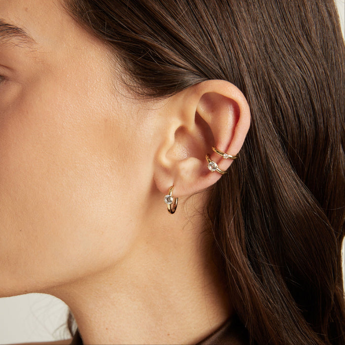 Model. Close up of woman wearing two gold ear cuffs and a huggie earring with white sapphires on neutral background.