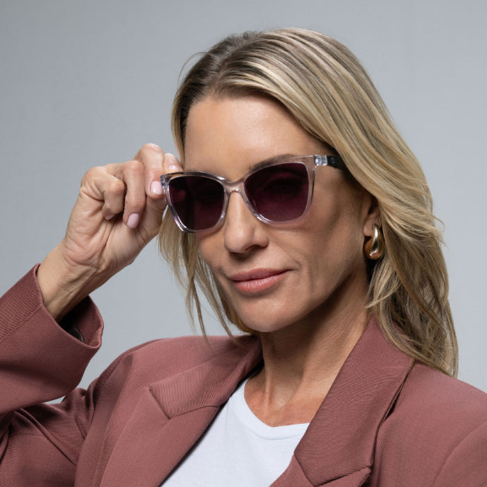 A blonde woman confidently wears Lucyd Moonrise champagne crystal sunglasses with polarized purple gradient lenses, paired with a mauve blazer, white shirt, and gold hoop earrings against a plain gray background.