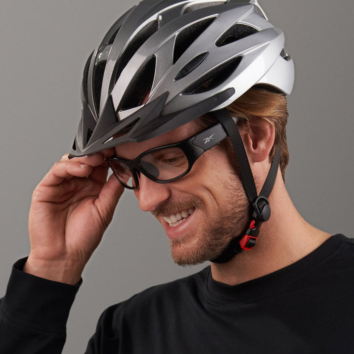 A smiling man wearing a black shirt, a silver bike helmet, and Reebok Nitrous Shift black glasses with adaptive photochromic lenses adjusts his glasses against a plain gray background.