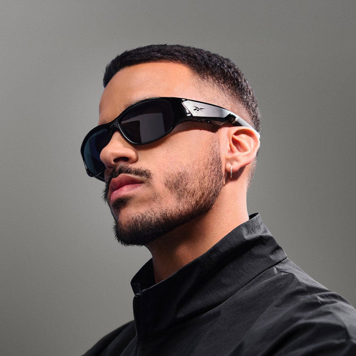 A man with short dark hair, a trimmed beard, and a small hoop earring wears Reebok Nitrous black polarized sunglasses and a black jacket, looking confidently to the side against a plain gray background.