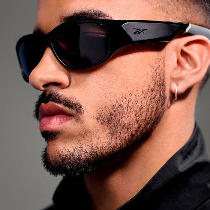 A close-up of a person with a trimmed beard and mustache wearing Reebok Nitrous sunglasses with polarized lenses, a small hoop earring, and a black collared shirt.