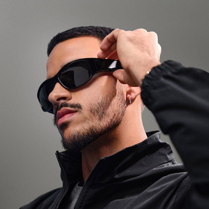 A man with a trimmed beard and mustache, wearing Reebok Nitrous black Bluetooth sport glasses and a black jacket, adjusts his glasses while looking to the side against a neutral background.