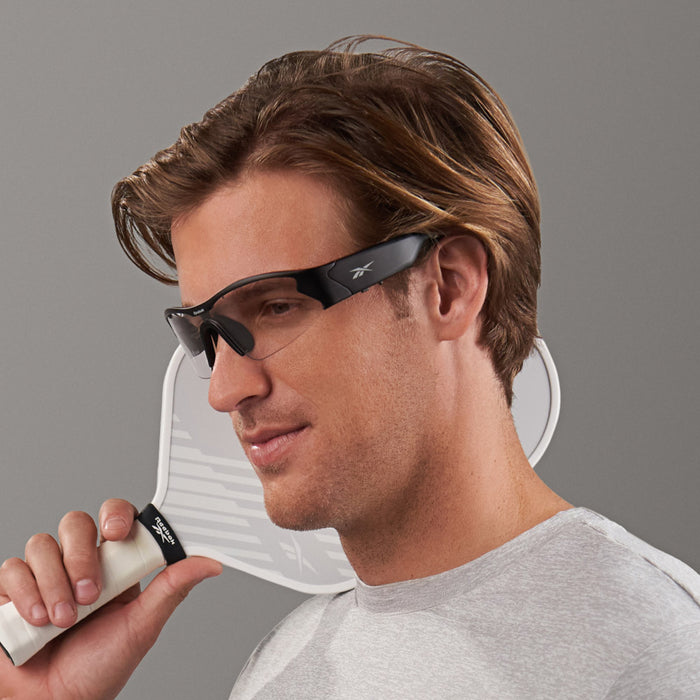 A man in a light gray shirt holds the Reebok Octane Shift, a white pickleball paddle, near his face while wearing black photochromic sports sunglasses against a plain gray background.