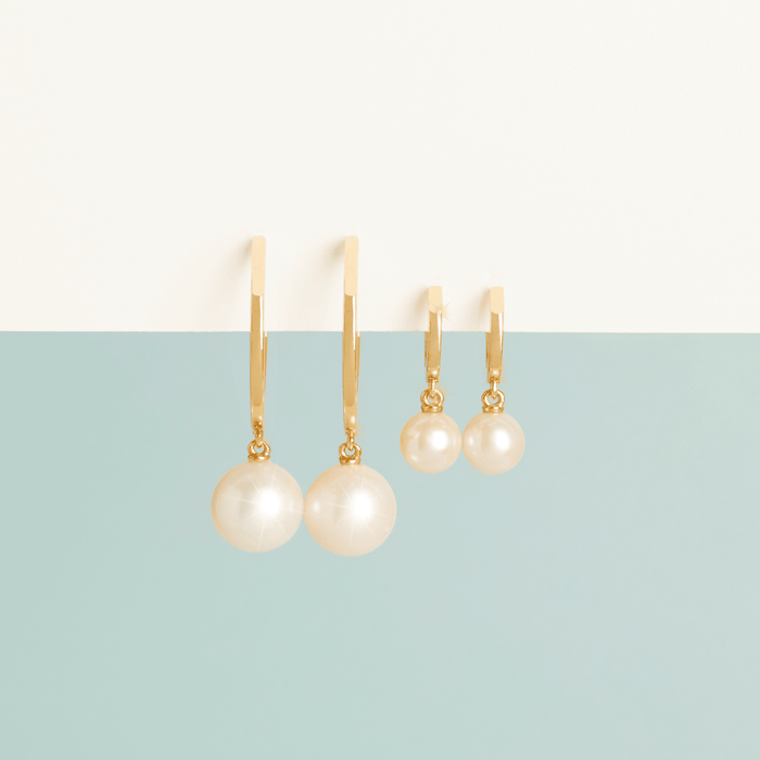 Flatlay Model Close-up of two pairs of gold earrings with dangling white pearls—one pair featuring long rectangular gold drops and the other featuring small huggie hoops—resting on a split white and light blue surface