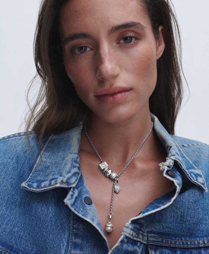 A close-up of a woman wearing a silver lariat-style necklace with a heart-shaped pendant and small cube charms, styled with a blue denim shirt against a light gray background.