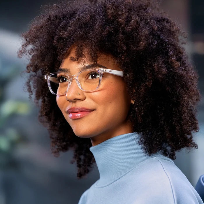 A woman with curly hair and clear Reebok Shine smart glasses—premium open-ear audio eyewear—smiles gently in a light blue turtleneck. The softly blurred background shows greenery and light.