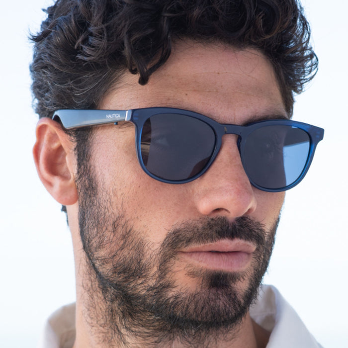 A man with short, curly dark hair and a beard wears Nautica Tailwind dark blue polarized UV400 glasses and a white collared shirt, glancing to the side against a light background.