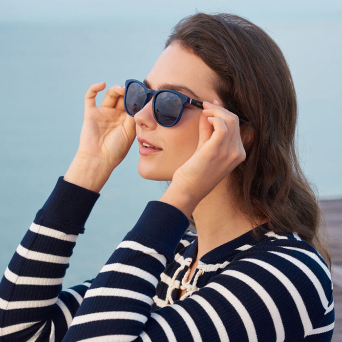 A woman with wavy brown hair wears Nautica Tailwind polarized UV400 sunglasses and a navy-and-white striped top, standing by the water and gazing into the distance on a sunny day.