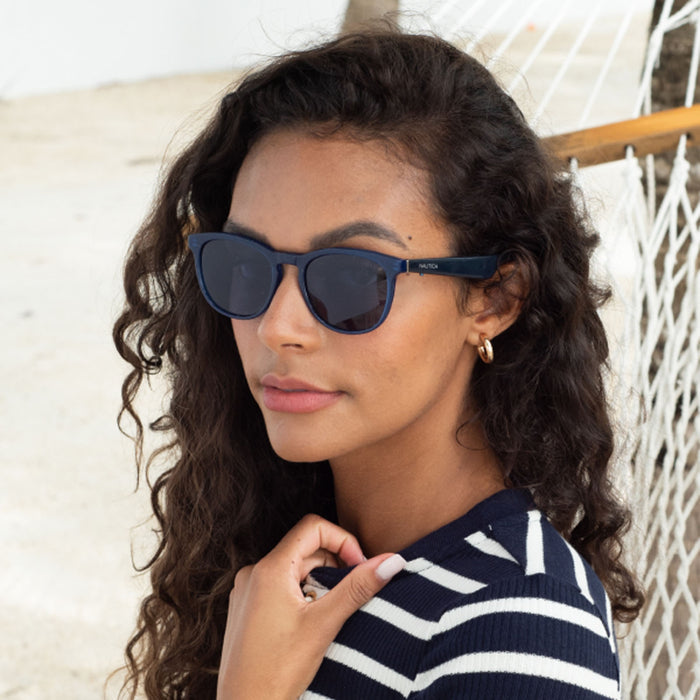 A woman with long curly hair, wearing Nautica Tailwind polarized UV400 glasses and a striped sweater, stands outdoors near a hammock, gazing calmly at the camera.