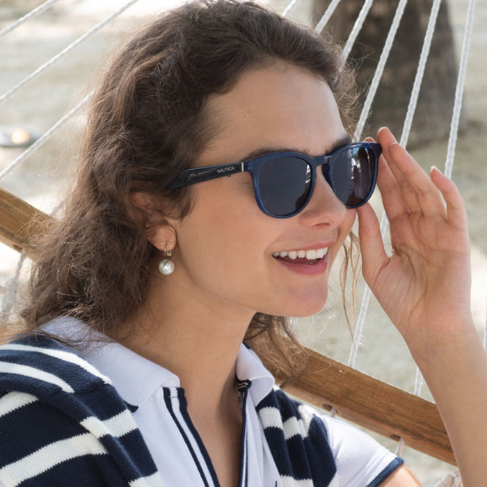 A woman with wavy brown hair wears Nautica Tailwind Bluetooth sunglasses with Lucyd patented temples, pearl earrings, and a navy and white striped top as she smiles on a hammock outdoors.