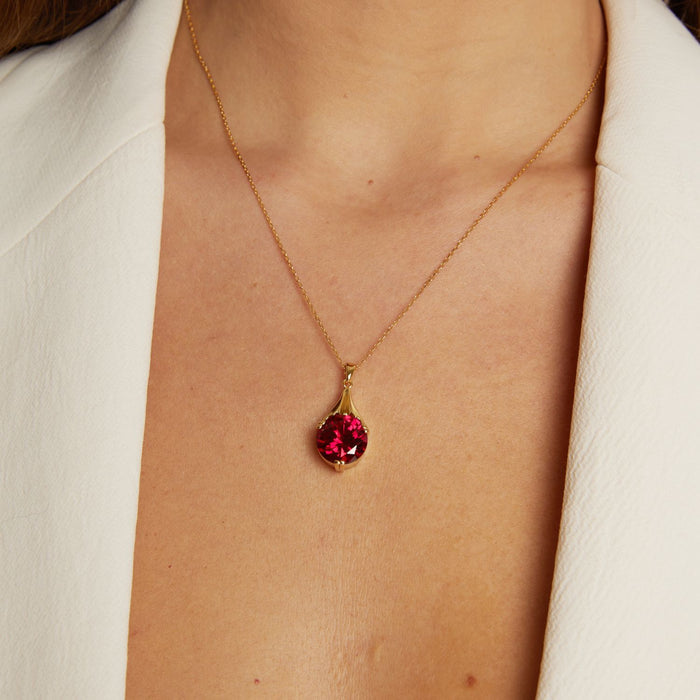 Model. Gold necklace with a red gemstone pendant on a person wearing a white shirt.