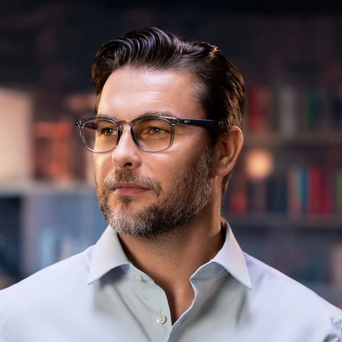 A man with short dark hair, a beard, and glasses, dressed in a light blue shirt, looks to the side while wearing Reebok Thunder smart glasses. Behind him are softly blurred bookshelves.